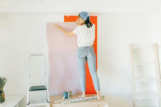 Frau bringt Fototapete an einer weißen Wand an – sie steht auf einem Tisch, trägt Jeans und ein gestreiftes Shirt; im Hintergrund sind eine Leiter, Farbrollen und weitere Tapetenrollen sichtbar.